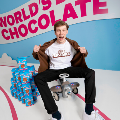Nolan riding a tricycle next to a stack of feastables chocolate boxes